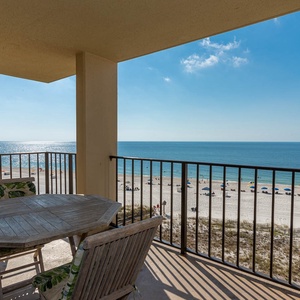 The balcony features a wooden table and chairs with a breathtaking view of the beach and sparkling water below