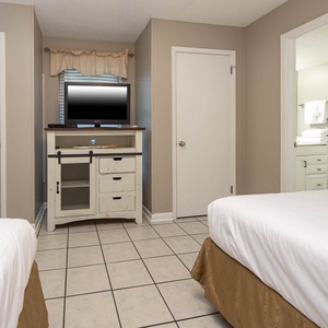 Bright guest room featuring two double beds, a TV on a rustic cabinet, and a doorway leading to a clean bathroom area