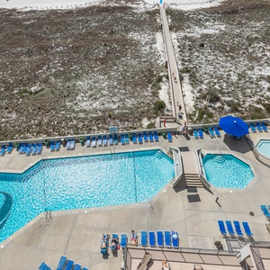 Aerial balcony view of the pool area featuring blue lounge chairs, umbrellas, and a pathway leading to the beach