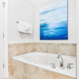 The master bath features a relaxing jetted tub, elegant beige tile, and a serene artwork, perfect for unwinding after a day