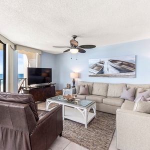 Bright living room with a large sectional sofa, a TV, and stunning views of the beach through expansive windows