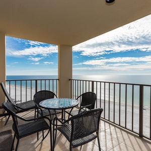 The balcony features a round table with mosaic design and four black wicker chairs, offering a serene view of the beach front