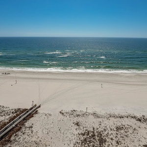 A panoramic view showcases the tranquil beach with soft white sand and gentle waves under a clear blue sky