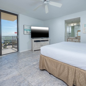 Bright master bedroom featuring a king bed, large TV, and a cozy turquoise chair by sliding glass doors leading to a balcony