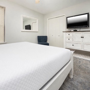 Elegant guest bedroom featuring a queen bed, a cozy blue chair, and a TV mounted above a white dresser