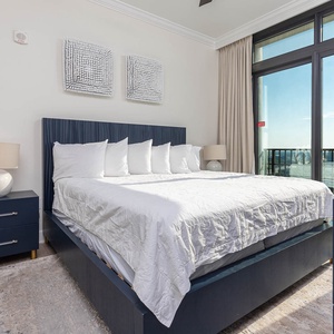 This bedroom features a stylish navy king bed, crisp white linens, and a stunning beachfront view through large windows