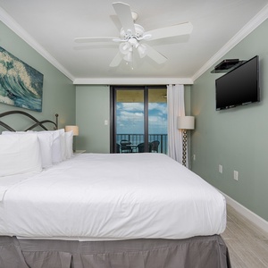 Bright master bedroom featuring a king bed, a wall-mounted TV, and the balcony with a view of the tranquil beachfront