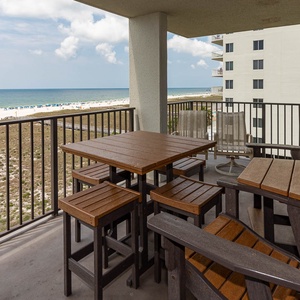 Balcony seating area features a wooden table and chairs, overlooking the beach with vibrant umbrellas dotting the shoreline