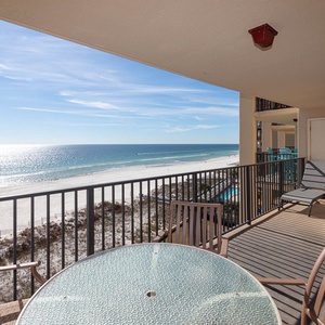 The balcony features a glass-top table and chairs, offering a serene view of the beach and sparkling water beyond