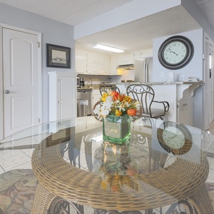 The dining area features a glass table with a floral centerpiece, leading to a well-equipped kitchen