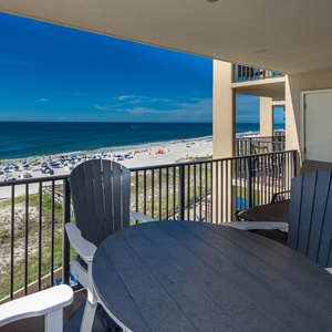 The balcony features a round table and chairs, offering a direct view of the beach with white sand and colorful umbrellas