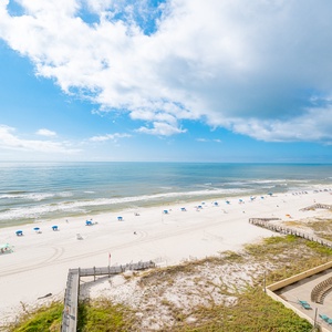 A serene view showcases a wide stretch of white sand beach lined with blue umbrellas, framed by gentle waves and a bright sky