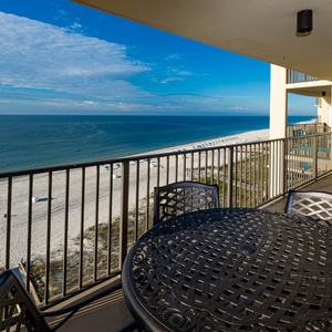 The balcony features a round table with chairs, offering a serene view of the white sand beach and tranquil waters