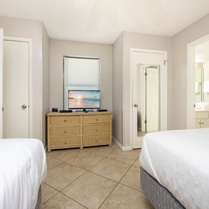 This guest bedroom features two double beds, a TV on a wicker dresser, and an adjoining bathroom