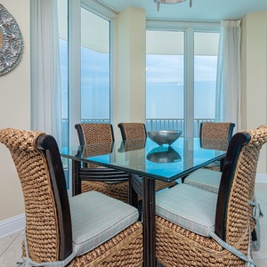 The dining area features a glass-top table surrounded by woven chairs, offering a stunning beach view through large windows
