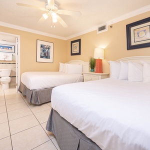 Bright guest bedroom featuring two double beds, a colorful lamp, and artwork, with a doorway leading to a bathroom area