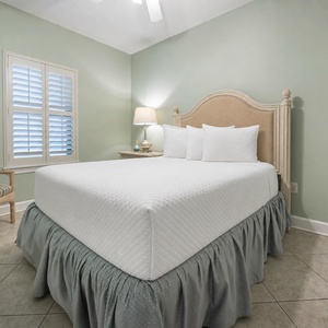 Serene guest bedroom featuring a plush queen bed, soft green walls, and a cozy chair beside a window with plantation shutters