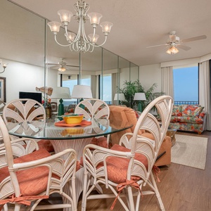 Dining area features a glass table surrounded by rattan chairs, with a vibrant view of the beach through large windows