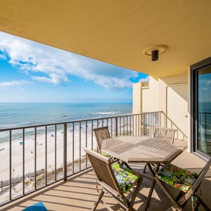 The balcony features a wooden table surrounded by chairs with floral cushions, overlooking a serene beach and calm waters