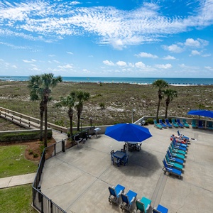 Inviting pool area features lounge chairs and a shaded table, with a view of the beach and glistening gulf waters beyond
