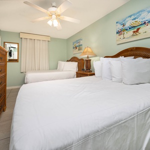 The inviting guest room features two double beds, a stylish wicker dresser, and beach-themed artwork, creating a relaxed vibe