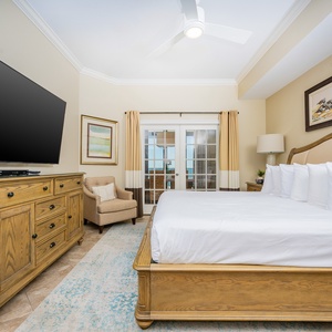 The master bedroom features a king bed, large TV, a cozy chair, and a view of the beach through sliding glass doors