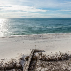 A wooden boardwalk leads to a tranquil beach with soft white sand and gentle waves under a bright, sunny sky