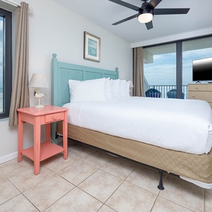 The master bedroom features a light blue headboard, a pink nightstand, and a TV, with a view of the beach from the window