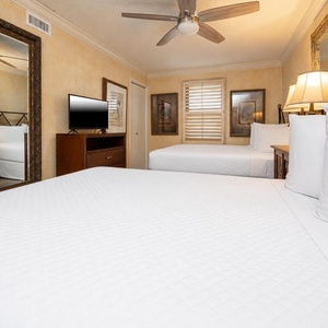 Bright guest bedroom featuring two double beds, a TV, decorative mirrors, and warm lighting for a welcoming atmosphere