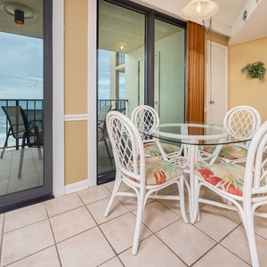 This dining area features a glass table with tropical-patterned chairs, offering a lovely view of the beach from the balcony