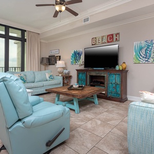 Relax in this living area featuring a large TV, turquoise accents, and expansive windows for a stunning beachfront view