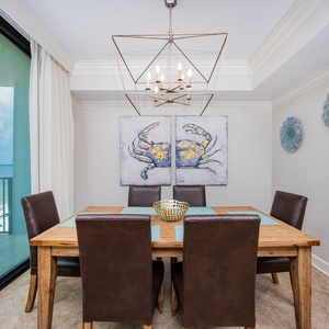 Dine at the wooden table with plush chairs, surrounded by beach-themed decor and a view of the inviting balcony