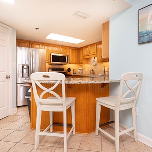 Kitchen area features wooden cabinetry, granite countertops, and a breakfast bar with two white stools for casual dining