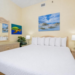 Guest bedroom features a large queen bed with white linens, a woven dresser, and coastal artwork on the walls