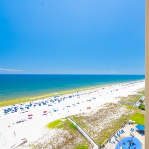 A balcony view showcases the beach and ocean, with colorful umbrellas dotting the white sand below under a clear blue sky