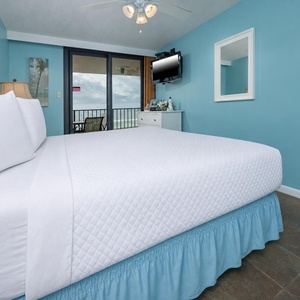 A bright master bedroom features a king bed with a blue skirt, a TV on the wall, and sliding doors leading to the balcony