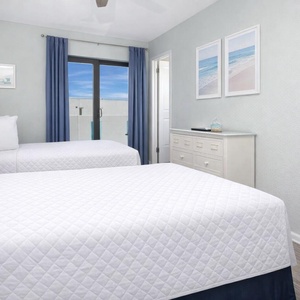This guest bedroom features two comfortable beds, light blue accents, and a view of the beach through large windows