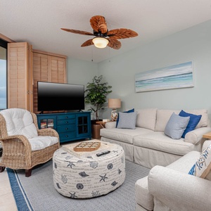 Cozy living area features a light sofa with queen sleeper, a wicker chair, and a view of the beach through large windows
