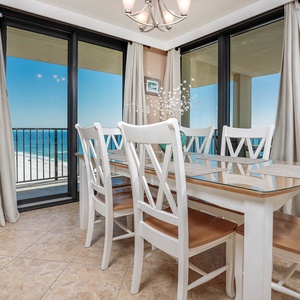 A bright dining area features a glass-top table with white chairs and offers a stunning view of the beach