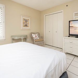 This bedroom featuring a cozy queen bed, a large TV, and charming wicker furniture against a soft beige backdrop