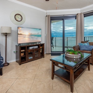 The living area features a large TV, comfortable seating, and access to a balcony with stunning views of the beach
