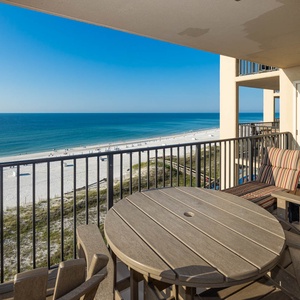 The balcony features a round table with chairs, offering a panoramic view of the beach and tranquil blue waters