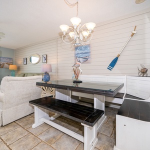 Bright dining area features a glass-top table with a stylish bench, surrounded by coastal decor and a view of the beach