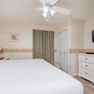 The bedroom features a king bed with crisp white linens, a flat-screen TV, and soft green curtains for a relaxing ambiance