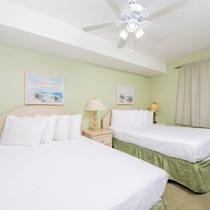 A guest room features two double beds with white linens, soft green walls, and beach artwork, creating a relaxed atmosphere