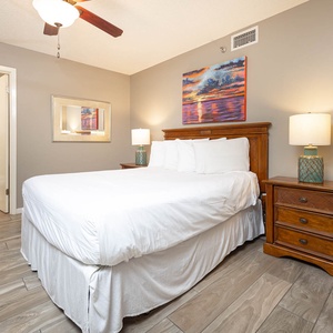 This cozy guest bedroom features a queen bed, wooden nightstand, and a vibrant sunset painting, with an adjoining bathroom