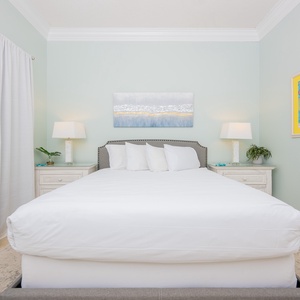Bright guest bedroom featuring a plush queen bed with crisp white linens, stylish lamps, and cheerful artwork on the wall