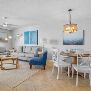 The living area features two gray sofas, a round dining table, and a view of the beach through large windows