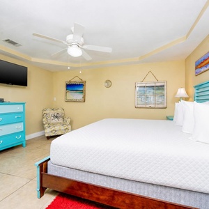 This cozy guest bedroom features a queen bed, vibrant turquoise furniture, a TV, and beach-themed decor for a relaxing stay