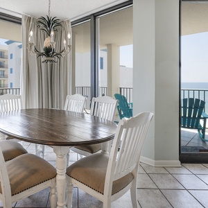 Bright dining area featuring a round wooden table and white chairs, with stunning views of the beach through large windows